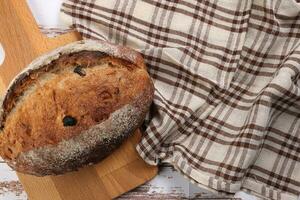 Rustic whole grain artisan bread loaf with cranberry raisin dry fruit nuts wrapped in checkers cloth with wooden chopping board over table top flat lay view photo