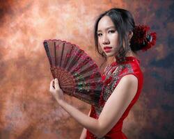 sur este asiático chino carrera étnico origen mujer vistiendo rojo terciopelo cheongsam con mano cosido secuencia trabajo vestir disfraz mano ventilador en retro Clásico antecedentes foto