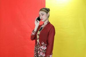 Asian woman traditional maroon kebaya sharong on red yellow paper background talk use phone smartphone photo