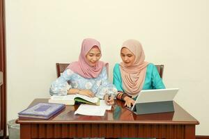 Two young Asian Malay Muslim woman wearing headscarf at home office student sitting at table talk mingle look at computer book document study discuss smile happy photo