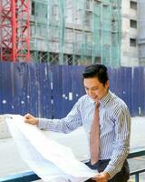 South East Asian young Malay Chinese man wearing formal tie looking at blueprint at outdoor construction site photo