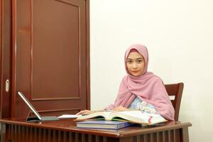 joven asiático malayo musulmán mujer vistiendo Pañuelo a hogar oficina estudiante sentar a mesa computadora leer libro archivo papel estudiar trabajo foto