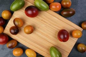 Red orange yellow green tomato mix variety on around wooden chopping board frame copy text space over black slate stone background photo