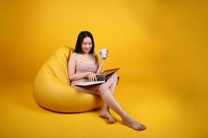 hermosa joven sur este asiático mujer sentar en un amarillo naranja bolsa de frijoles asiento color antecedentes relajarse leer estudiar pensar libro revista sostener blanco café taza foto