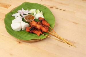 Chicken beef mutton meet Satay set nasi impit sticky rice cake sliced onion cucumber sweet peanut sauce on banana leaf bamboo basket on wooden background photo