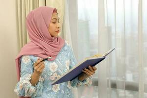 young Asian Malay Muslim woman wearing headscarf at home office next to window look at book document study think write photo