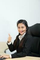 Young Asian female wearing suit sitting at office desk look at camera headphone call center thumbs up photo