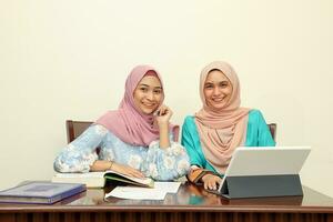 Two young Asian Malay Muslim woman wearing headscarf at home office student sitting at table talk mingle look at computer book document study discuss smile happy photo
