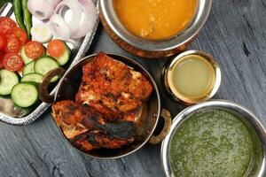 Spicy red Chicken grilled tikka tandoori nan bread green spinach curry yogurt sauce dal tomato cucumber onion salad set in metal stainless steel brass copper pot on black char wood background photo