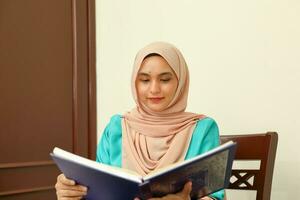 young Asian Malay Muslim woman wearing headscarf at home office student sitting at table read book document note study smile happy look photo