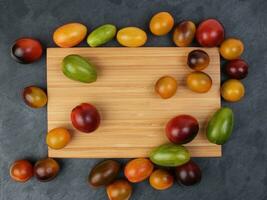 Red orange yellow green tomato mix variety on around wooden chopping board frame copy text space over black slate stone background photo