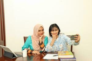 Two young Asian Malay Muslim woman wearing headscarf at home office student sitting at table phone computer book document selfie self portrait with smartphone photo