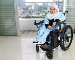 Young woman south east Asian malay wearing headscarf sitting on wheelchair against window light. Paralyzed waist down from birth. photo