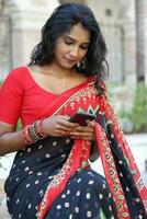 joven atractivo hermosa indio mujer en rojo y negro sari mensajes de texto leyendo masaje teléfono inteligente al aire libre urbano ladrillo pared antecedentes foto