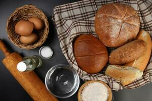 Freshly baked bread loaf bun roll round long mix verity wrapped in checkered kitchen fabric napkin towel wheat flower oil water salt eggs rolling pin slate stone over black background photo