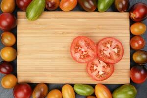 Red orange yellow green tomato mix variety on around wooden chopping board frame copy text space over black slate stone background photo