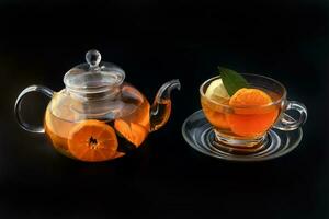 Liquid tea lemon orange slice green leaf cinnamon stick in transparent glass teacup saucer teapot kettle on black background photo