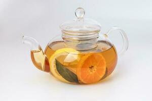 Liquid tea lemon orange slice green leaf cinnamon stick in transparent glass teapot kettle on white background photo