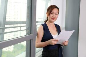 beautiful attractive middle age southeast asian woman home office work read see document standing near glass window look at camera urban daylight window background photo