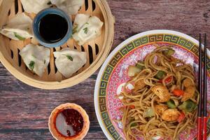 Home style dim sum dumpling in bamboo steamer chili flake oil sauce Stair fried noodle with prawn seafood tofu colorful pepper on oriental style plate chopsticks over rustic wooden background photo