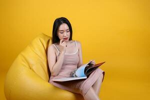 Beautiful young south east Asian woman sit on a yellow orange beanbag seat color background relax read study think book magazine photo
