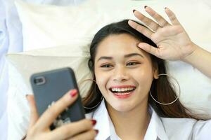 Young beautiful modern Malay woman lying on white pillow bed wearing white shirt using smartphone video chat talk smile wave selfie photo