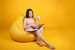 hermosa joven sur este asiático mujer sentar en un amarillo naranja bolsa de frijoles asiento color antecedentes relajarse leer estudiar pensar libro revista sostener blanco café taza foto