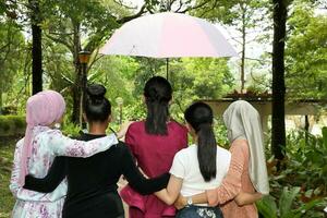 group of woman friends Malay Chinese Indian Asian outdoor park  nature walking under one umbrella secure safe unity hands on shoulder and waist photo