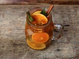 Liquid ice lemon orange tea with slice green leaf cinnamon stick in transparent glass jar mug on rustic wood background whole fruit photo