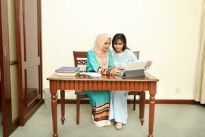 Two young Asian Malay Muslim woman wearing headscarf at home office student sitting at table talk mingle look at computer book document study discuss read type point show photo