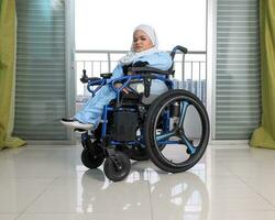 Young woman south east Asian malay wearing headscarf sitting on wheelchair against window light. Paralyzed waist down from birth. photo