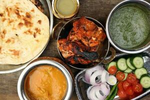 Spicy red Chicken grilled tikka tandoori nan bread green spinach curry yogurt sauce dal tomato cucumber onion salad set in metal stainless steel brass copper pot on rustic vintage wood background photo