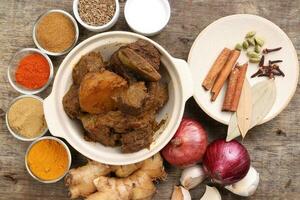Beef bhuna dry curry with potato in ceramic bowl ingredients chilli cumin coriander turmeric powered salt onion garlic ginger bay leaf cinnamon cardamom clove over rustic wood background photo