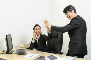 Young Asian male female wearing suit sitting at office desk joy high five happy success photo