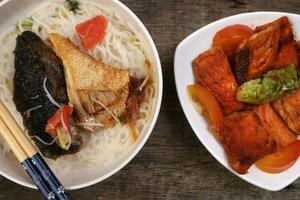 Fish head noodles vermicelli milk soup in white bowl pan friend salmon fish fillet on triangular plate chopstick spoon on rustic wooden background photo