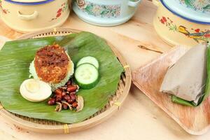 Nasi Lemak fragrant rice cooked in coconut milk served with sambal friend peanut anchovy boiled egg packed in banana leaf on round bamboo plate on wooden background photo
