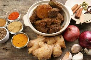 Beef bhuna dry curry with potato in ceramic bowl ingredients chilli cumin coriander turmeric powered salt onion garlic ginger bay leaf cinnamon cardamom clove over rustic wood background photo