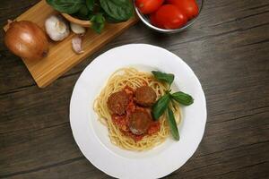 Cooked spaghetti sauce meat ball cheese powder on white plate with ingredients roma tomato glass bowl basil onion garlic wooden board recipe on table marble wall photo