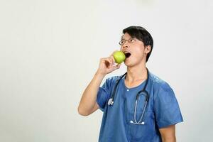 Young Asian male doctor wearing apron uniform tunic stethoscope holding pointing showing eating healthy green apple photo