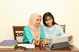 Two young Asian Malay Muslim woman wearing headscarf at home office student sitting at table talk mingle look at computer book document study discuss read type point show photo