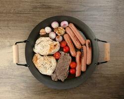 Mix meet pan chicken breast beef steak sausage tomato onion garlic oil fry grill on metal frying pan two wood handle on rustic wooden background photo