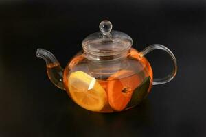 Liquid tea lemon orange slice green leaf cinnamon stick in transparent glass teapot kettle on black background photo