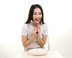 Beautiful young south east Asian woman pretend acting posing holding empty fork spoon in hand eat taste look see white plate on table white background smile happy exited photo