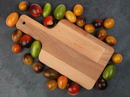 Red orange yellow green tomato mix variety on around wooden chopping board frame copy text space over black slate stone background photo