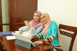 Two young Asian Malay Muslim woman wearing headscarf at home office student sitting at table talk mingle look at computer book document study discuss smile happy photo