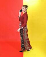 Asian woman traditional maroon kebaya sharong on red yellow paper background talk use phone smartphone photo