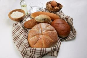 Freshly baked bread loaf bun roll round long mix verity wrapped in checkered kitchen fabric napkin towel wheat flower oil water salt eggs over white background photo