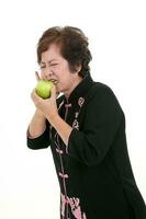 Elderly asian Chinese female wearing black tang samfu on white background eating biting green apple tooth ace pain photo