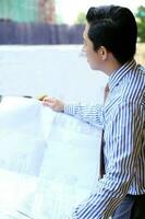 South East Asian young Malay Chinese man wearing formal tie looking at blueprint at outdoor construction site photo