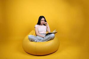 hermosa joven sur este asiático mujer se sienta en un amarillo bolsa de frijoles asiento naranja amarillo color antecedentes actitud Moda estilo elegante belleza estado animico expresión descanso relajarse leer libro pensar foto